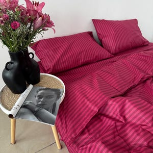 May include: A bedding set featuring a deep magenta and dark pink striped pattern, including a duvet cover and two pillowcases. A black vase with pink lilies and a magazine are placed on a small round table next to the bed.