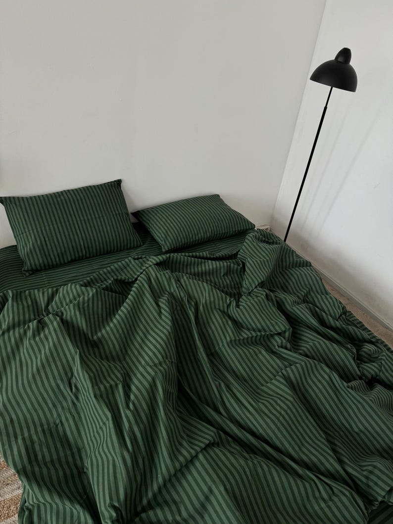 Puede incluir: Una s&aacute;bana de cama a rayas verdes con fundas de almohada a juego. La cama est&aacute; hecha con la s&aacute;bana y las almohadas, y el edred&oacute;n est&aacute; doblado al pie de la cama.