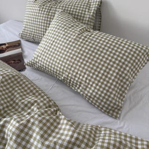 Pode incluir: Um conjunto de roupa de cama com um padrão de xadrez vichy em verde sálvia e branco. O conjunto inclui duas fronhas retangulares e uma capa de edredão a condizer, exibindo o design de xadrez pequeno em toda a superfície do tecido.