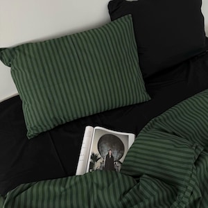 Puede incluir: Una almohada a rayas verdes con una revista en blanco y negro encima. La almohada está sobre una cama negra con una manta a rayas verdes.