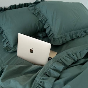 Könnte beinhalten: Ein silberner Laptop steht auf einem Bett mit grüner, gekräuselter Bettwäsche. Der Laptop ist geöffnet und das Apple-Logo ist auf dem Deckel sichtbar.