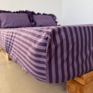 Può includere: Un lenzuolo con angoli e due federe abbinate caratterizzate da un motivo a righe verticali in tonalità alternate di viola scuro e lavanda chiaro. Le federe sono rifinite con un dettaglio a volant lungo il perimetro.