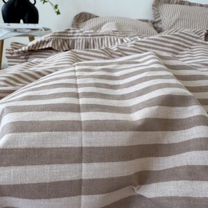 May include: Close-up of a beige and white striped bedding set. The set includes a duvet cover and pillowcases. The fabric has a soft, textured appearance. The bedding is arranged on a bed, with a vase and greenery in the background.