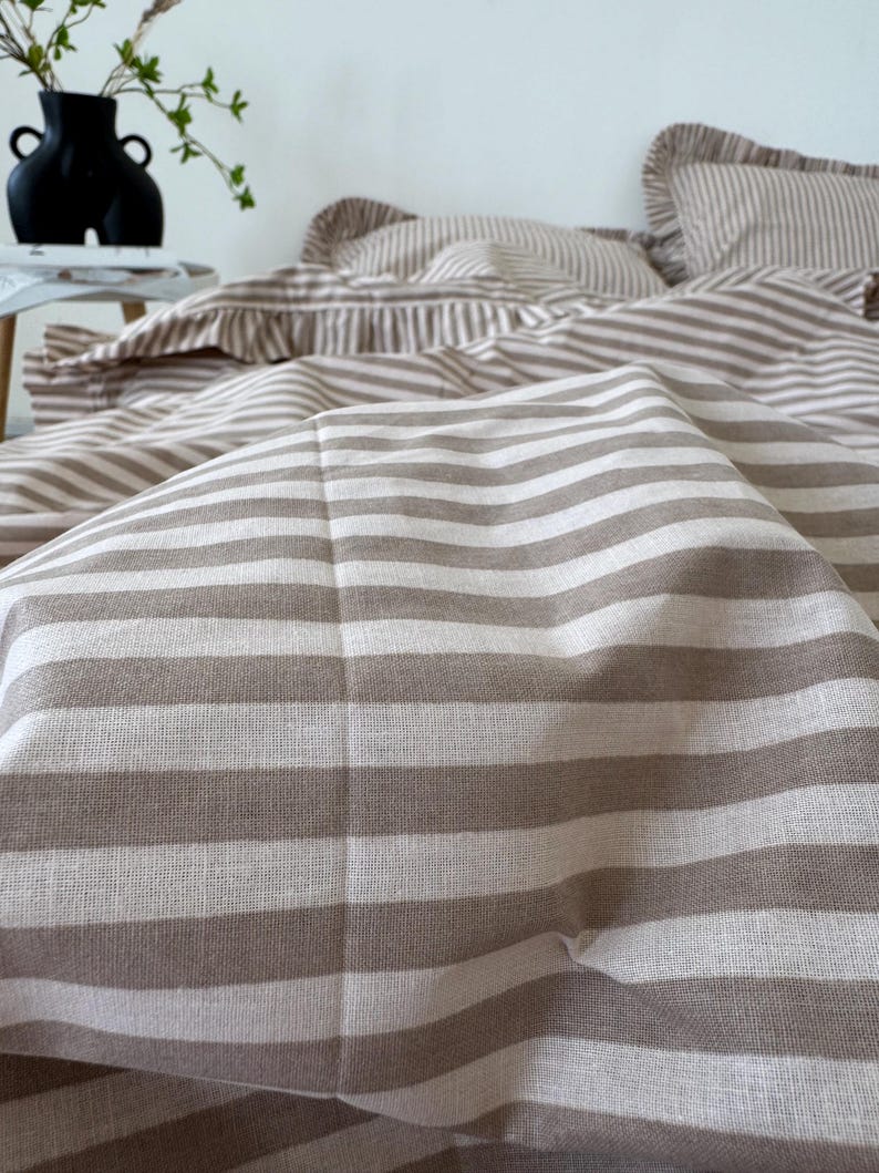 May include: Close-up of a beige and white striped bedding set. The set includes a duvet cover and pillowcases. The fabric has a soft, textured appearance. The bedding is arranged on a bed, with a vase and greenery in the background.