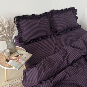 Könnte beinhalten: Ein dunkelviolettes gestreiftes Bettwäsche-Set, bestehend aus einem Bettbezug und zwei Kissenbezügen mit Rüschenkanten. Die Bettwäsche ist auf einem Bett drapiert, mit einer Zeitschrift und dekorativen Gegenständen auf einem Beistelltisch. Thema: Schlafzimmerdekoration.