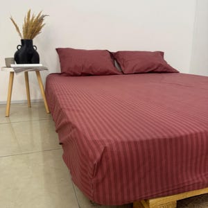 May include: A bed with a burgundy striped sheet set and two matching pillowcases. The bed is on a wooden pallet. A small side table with a black vase and dried plants is to the left.
