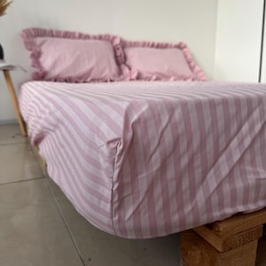 May include: A bed set featuring a light pink and white striped fitted sheet and two matching pillowcases with ruffled edges. The bedding is arranged on a low platform base, showcasing the soft pastel color palette and the vertical striped pattern.