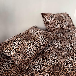Puede incluir: Un juego de cama con un estampado de leopardo en tonos marrón, negro y beige. El conjunto incluye una funda de almohada rectangular y una sábana o funda nórdica a juego, ambas cubiertas con un diseño de piel animal con manchas oscuras.