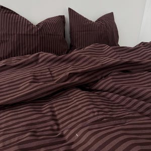 May include: A bed set featuring a duvet cover and two pillowcases in a deep burgundy color with a striped pattern. The bedding is made of a soft, wrinkle-resistant fabric, creating a cozy and inviting bedroom aesthetic.