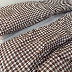 Pode incluir: Um conjunto de roupa de cama com um padrão de xadrez vichy castanho e branco. O conjunto inclui duas almofadas retangulares e uma capa de edredão a condizer, exibindo todas o mesmo padrão de xadrez pequeno em toda a superfície.