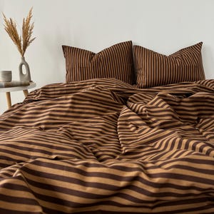 May include: A brown and tan striped bed set, including a duvet cover and two pillowcases. The bedding is arranged on a bed, with a white wall in the background. A small table with a vase and decorative items is visible in the image.