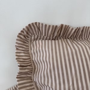 May include: Close-up of a striped pillowcase with a ruffled edge. The pillowcase features alternating vertical stripes in a light brown and off-white color scheme. The ruffled edge adds a decorative touch to the bedding.