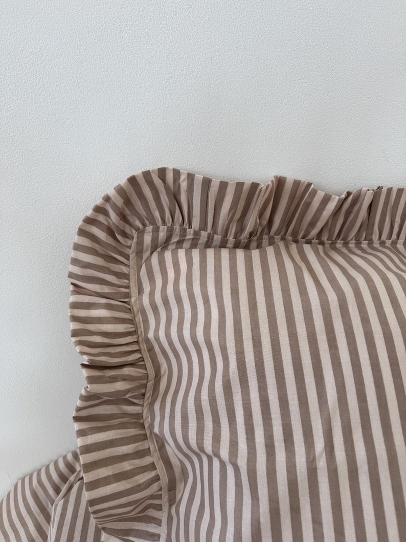 May include: Close-up of a striped pillowcase with a ruffled edge. The pillowcase features alternating vertical stripes in a light brown and off-white color scheme. The ruffled edge adds a decorative touch to the bedding.