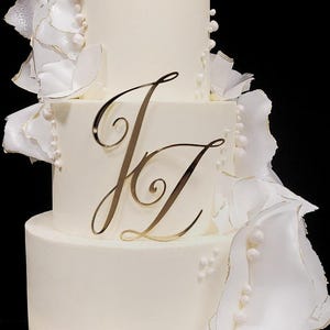 Può includere: Una torta nuziale bianca a tre piani decorata con fiori di zucchero bianchi e dettagli di perle. La torta è sormontata da un monogramma dorato "J & Z".