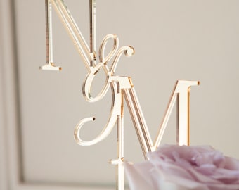 May include: A gold-toned acrylic cake topper featuring the capital letters N and M connected by an ampersand symbol. The letters are rendered in a clean, serif typeface with a reflective surface, positioned upright above a cake decorated with light purple roses.