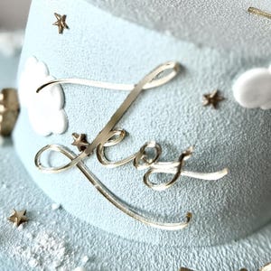 Op de afbeelding: Een lichtblauwe cake met glazuur, versierd met twee roze teddybeerfiguren met gouden feesthoeden. De cake heeft gouden accenten, witte wolken- en sterdecoraties en het cijfer "1". De naam "Leo" is in gouden letters geschreven.