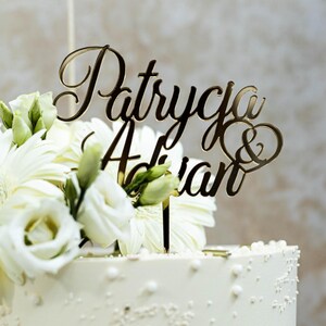 May include: A gold-colored acrylic cake topper featuring the names Patrycja & Adrian in a cursive script font, with an ampersand enclosed in a heart shape. The topper is inserted into a white cake decorated with white flowers and small white beads.