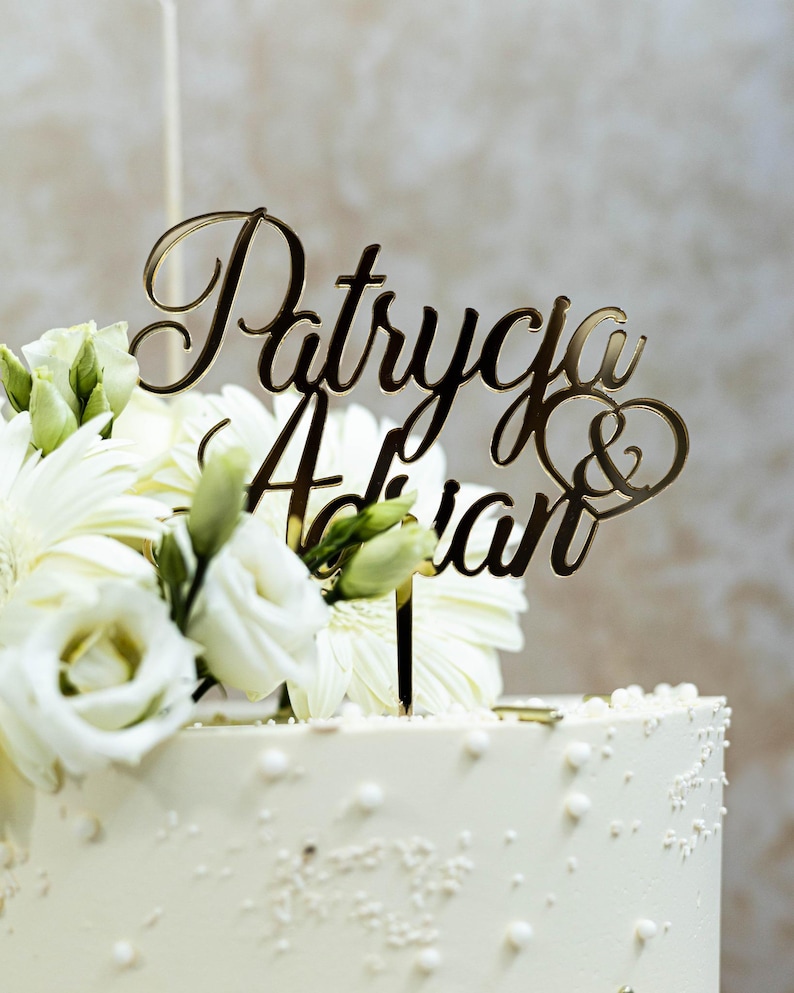 May include: A gold-colored acrylic cake topper featuring the names Patrycja & Adrian in a cursive script font, with an ampersand enclosed in a heart shape. The topper is inserted into a white cake decorated with white flowers and small white beads.