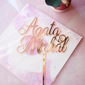 May include: A rose gold mirrored acrylic cake topper featuring the names Agata and Michal in a cursive script font, with a small heart symbol containing an ampersand positioned between the names, attached to a single vertical support stake.