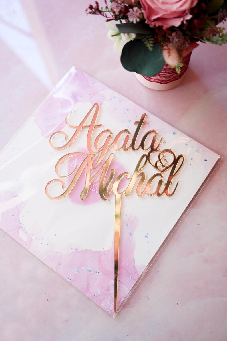 May include: A rose gold mirrored acrylic cake topper featuring the names Agata and Michal in a cursive script font, with a small heart symbol containing an ampersand positioned between the names, attached to a single vertical support stake.