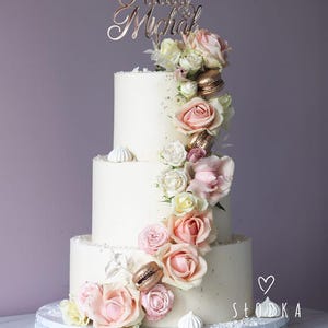 May include: A three-tier white cake decorated with pink and cream roses, gold macarons, and small white meringues. A rose gold cake topper features the names Paula and Michał. The text Słodka Fabryka with a heart icon is visible in the corner.
