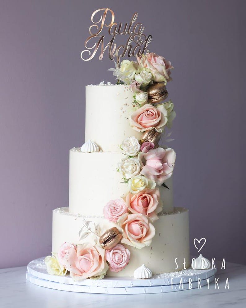 May include: A three-tier white cake decorated with pink and cream roses, gold macarons, and small white meringues. A rose gold cake topper features the names Paula and Michał. The text Słodka Fabryka with a heart icon is visible in the corner.
