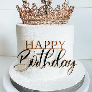 Könnte beinhalten: Ein weißer Kuchen mit Zuckerguss, gekrönt von einer goldenen Krone und einem goldenen "Happy Birthday" Tortenaufsatz. Die Krone ist mit klaren Kristallen verziert. Der Kuchen steht auf einem weißen Kuchenständer.