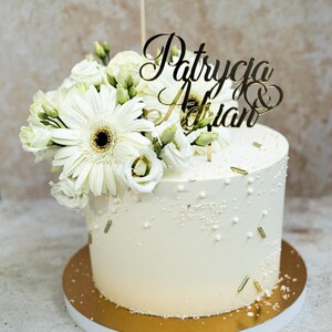 May include: A cylindrical white cake decorated with white and gold sprinkles, topped with a cluster of white gerbera daisies and lisianthus flowers, and a gold-colored acrylic cake topper featuring the names Patrycja & Adrian in script.