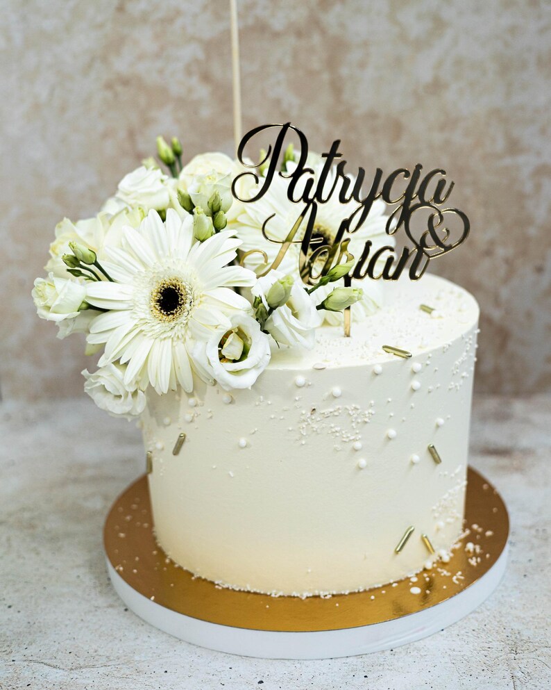 May include: A cylindrical white cake decorated with white and gold sprinkles, topped with a cluster of white gerbera daisies and lisianthus flowers, and a gold-colored acrylic cake topper featuring the names Patrycja & Adrian in script.
