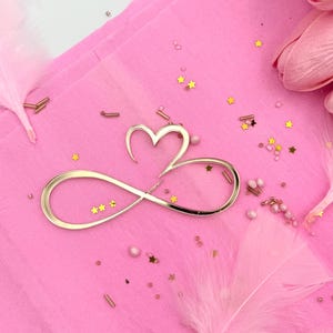 May include: A gold-coloured infinity symbol with a heart shape at the top, decorated with three small gold stars. The symbol sits on a pink surface with scattered gold star-shaped confetti, small pearls, and pink feathers.