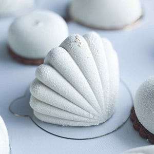 Puede incluir: Primer plano de varios pasteles blancos. El principal tiene forma de concha, con una superficie texturizada y helada. Otros pasteles son redondos o en forma de cúpula. Algunos tienen un acento de hoja de oro. Los pasteles están sobre pequeñas bases redondas.