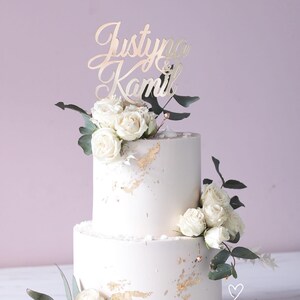 May include: A two-tier cake with smooth light-colored frosting, decorated with gold leaf accents, white roses, and green eucalyptus leaves. A gold-colored script cake topper reads Justyna & Kamil. A small heart and the text SŁODKA FABRYKA are visible.