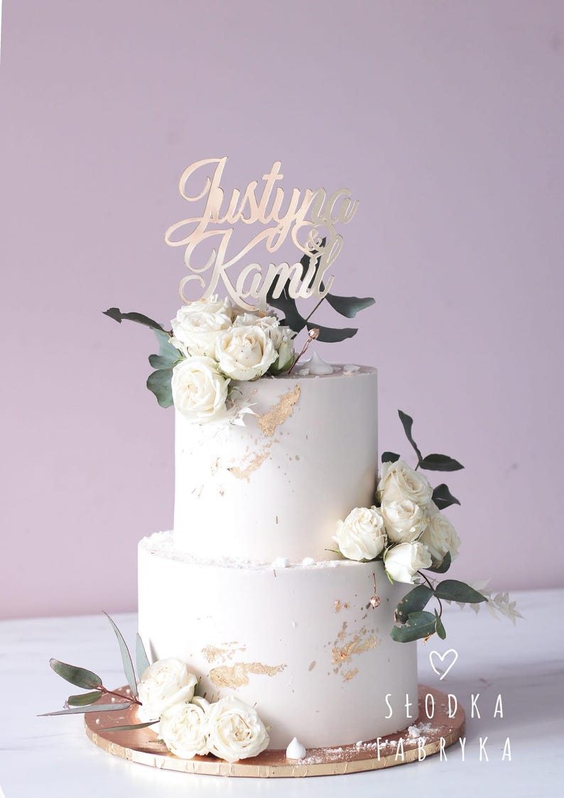 May include: A two-tier cake with smooth light-colored frosting, decorated with gold leaf accents, white roses, and green eucalyptus leaves. A gold-colored script cake topper reads Justyna & Kamil. A small heart and the text SŁODKA FABRYKA are visible.