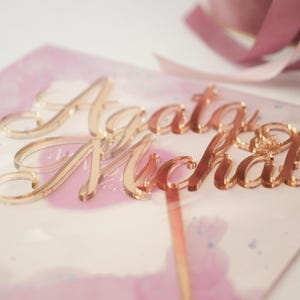 May include: A rose gold acrylic cake topper featuring the names Agata and Michal in a cursive script font. The topper is placed on a light pink watercolor card with a thin rose gold stem extending downwards from the lettering.