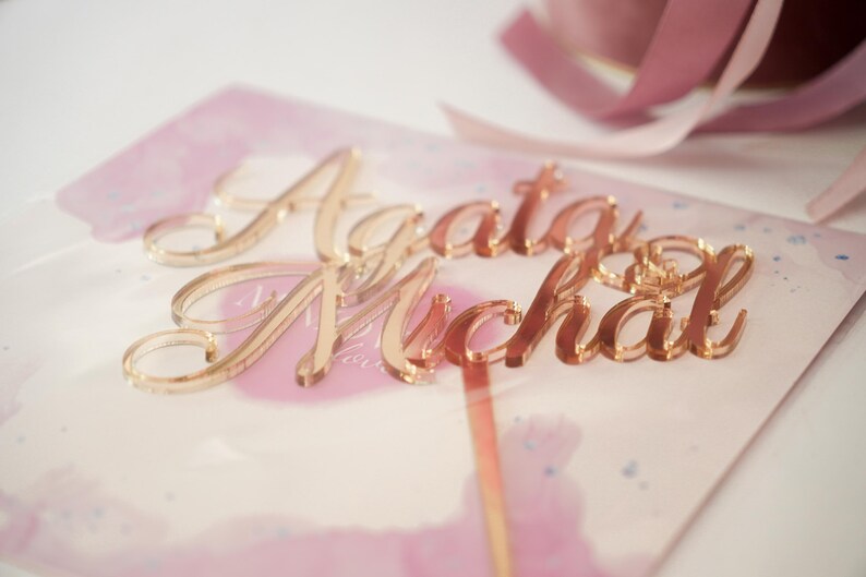 May include: A rose gold acrylic cake topper featuring the names Agata and Michal in a cursive script font. The topper is placed on a light pink watercolor card with a thin rose gold stem extending downwards from the lettering.