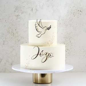 Può includere: Una torta bianca a due piani decorata con una colomba marrone che porta un ramo, il nome "Józio" scritto in corsivo marrone e dettagli in foglia d'oro.