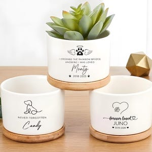 May include: Three white ceramic planters with wooden bases. One planter holds a succulent. The planters are decorated with pet memorial designs and the names "Monty", "Candy", and "Juno". The text includes dates and the phrase "Never Forgotten".