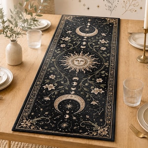 May include: A rectangular black table runner featuring a gold and white celestial print with a central sun face, moon phases, stars, and floral vines. The runner is centered on a light wood dining table set with white plates, glasses, and candles.