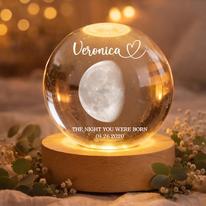 May include: A clear glass sphere with a moon phase design, illuminated from a wooden base. The sphere has the name "Veronica" and the text "THE NIGHT YOU WERE BORN 04.26.2020". Decorative sprigs of greenery and flowers surround the base.