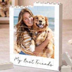 May include: A white brick-style photo frame with a picture of a woman and a golden retriever dog. The words "My Best Friends" are written in a script font. Additional white bricks are scattered around the frame. A framed photo of a puppy is in the background.