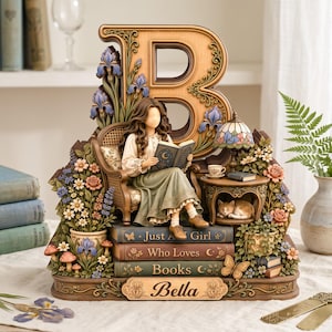 May include: A decorative figurine featuring a large wooden letter B, a person sitting in a chair reading a book, and a sleeping cat under a side table. The base includes stacked books labelled Just A Girl Who Loves Books and a nameplate reading Bella.
