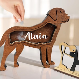 May include: A wooden dog-shaped keepsake box with the name "Alain" in white script. The box is a dark brown color and has a cutout area to hold pet fur. A small wooden dog-themed decorative piece is also present.