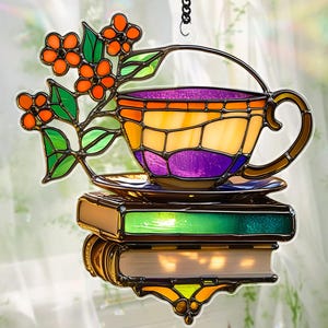 May include: Stained glass suncatcher featuring a teacup with a handle, resting on a stack of books. The teacup is filled with purple liquid and has orange, yellow, and purple panels. Orange berries and green leaves adorn the top.