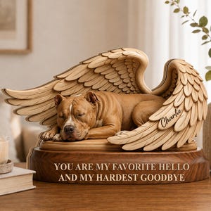 May include: A wooden memorial sculpture featuring a brown dog sleeping peacefully between two large, detailed angel wings. The base is engraved with the text, "YOU ARE MY FAVORITE HELLO AND MY HARDEST GOODBYE," and the name "Charlie" is etched on one wing.