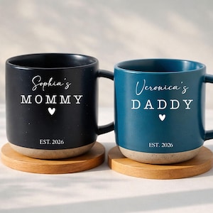 May include: Two ceramic mugs, one black and one teal, each with a wooden coaster. The black mug says "Sophia's MOMMY" and the teal mug says "Veronica's DADDY" in white script. Both mugs have a white heart and "EST. 2026".