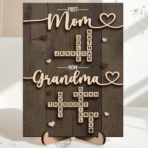 May include: Wooden sign with the words "First Mom" and "Now Grandma" in a cursive font. The sign has a dark wood grain background with names spelled out in a crossword puzzle style. Small heart and paw print accents are included.