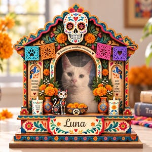 May include: A wooden pet memorial frame featuring a central photo of a white cat, topped with a sugar skull. The frame is decorated with colorful floral patterns, papel picado banners, and the name Luna. It includes miniature cat figurines and candles.