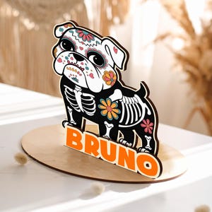May include: A wooden bulldog figurine with a Day of the Dead design. The dog is black with a white head and floral accents. The word "BRUNO" is displayed in orange block letters on a wooden base.