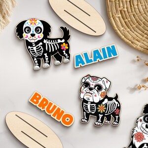 May include: Wooden dog-shaped ornaments with Day of the Dead skeleton designs. The dogs have colorful floral accents and are paired with name tags that read "ALAIN" and "BRUNO". Decorative wooden surfboard shapes are also included.