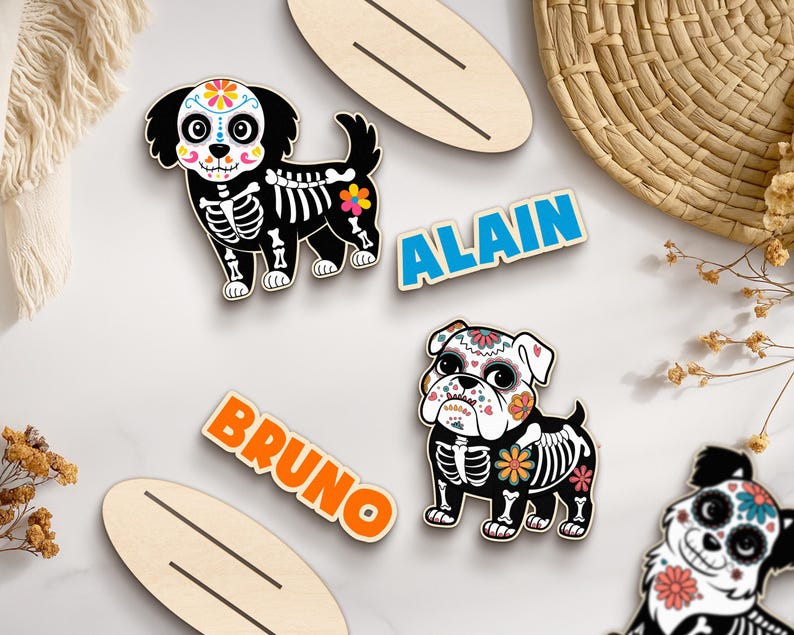 May include: Wooden dog-shaped ornaments with Day of the Dead skeleton designs. The dogs have colorful floral accents and are paired with name tags that read "ALAIN" and "BRUNO". Decorative wooden surfboard shapes are also included.